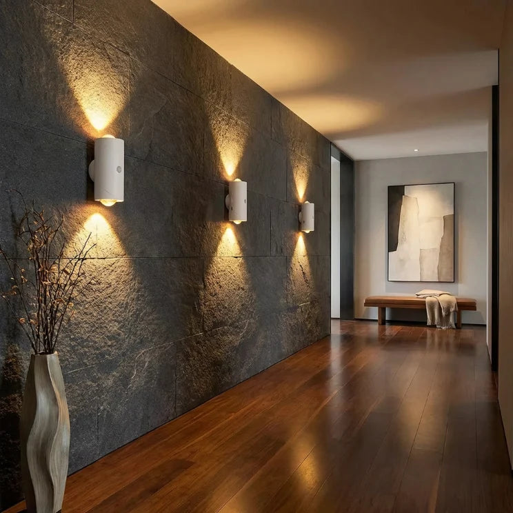 Couloir moderne éclairé par trois appliques murales sans fil design projetant un faisceau lumineux haut et bas sur un mur en pierre sombre.