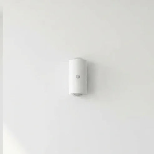 Applique murale sans fil blanche et cylindrique avec capteur de mouvement central, fixée sur un mur clair.