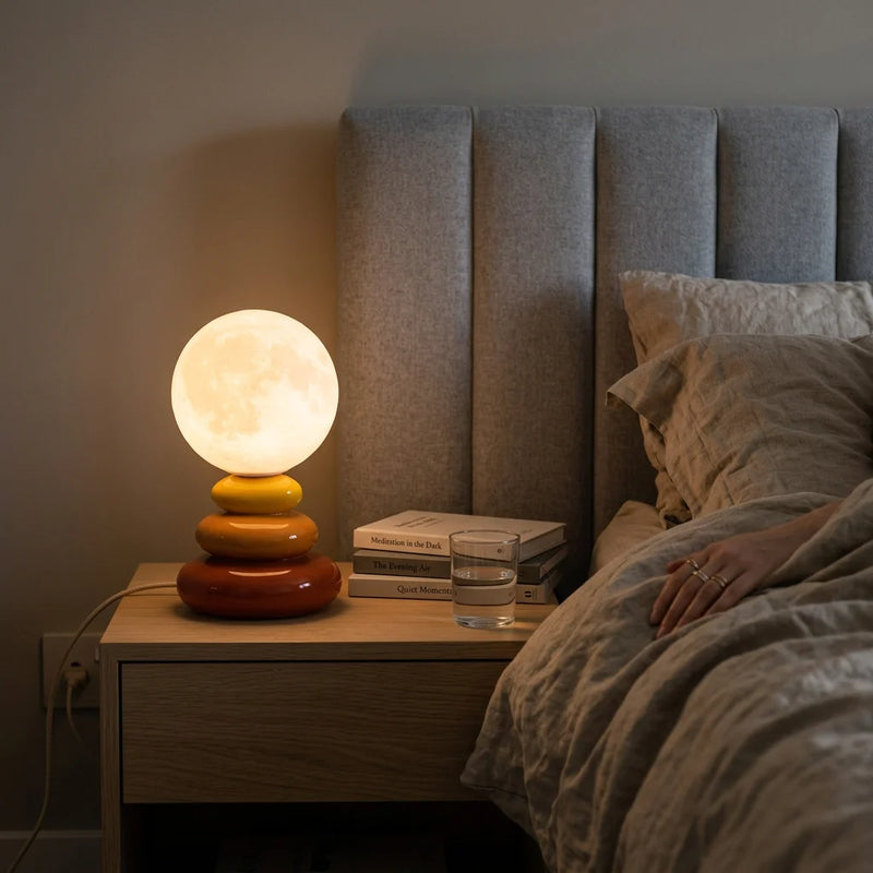 Lampe de chevet céramique lune LED orange allumée sur table de nuit en bois, ambiance chambre adulte avec lumière ambrée douce filtrée céramique et draps en lin froissé — Tendre Veilleuse