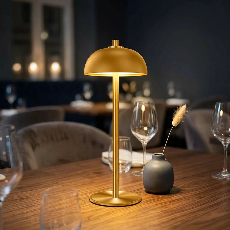 Lampe champignon dorée sans fil allumée en lumière chaude sur
table de restaurant en bois sombre, ambiance lounge avec
éléments décoratifs floutés — Tendre Veilleuse