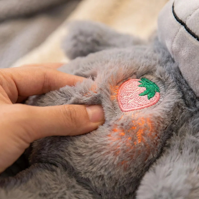 Gros plan sur une main touchant la fourrure grise d'une peluche sensorielle avec une broderie fraise rose sur le ventre.
