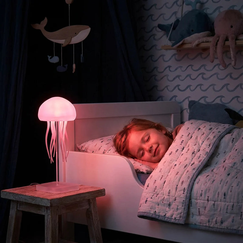 Enfant dormant paisiblement dans un lit entouré d'une décoration océanique, éclairé par la lueur rose douce d'une veilleuse méduse sur la table de chevet pour calmer la peur du noir.