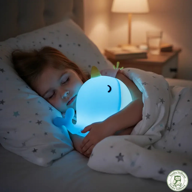 Enfant dormant paisiblement dans son lit en serrant contre lui sa veilleuse narval bleue qui diffuse une lumière douce