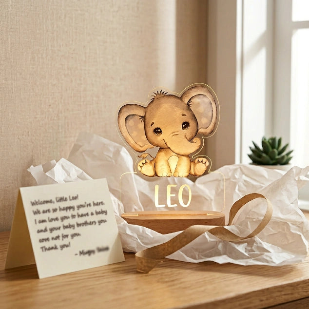 Veilleuse personnalisée prénom animal présentée comme cadeau de naissance, entourée de papier de soie blanc et ruban kraft naturel, lumière allumée ambrée — Tendre Veilleuse