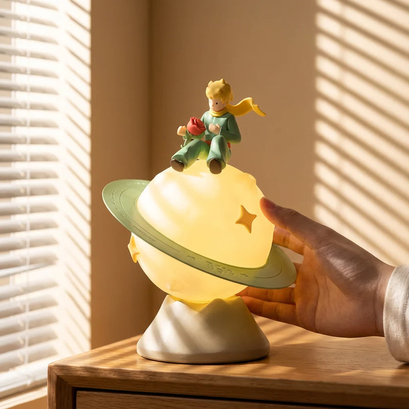 Une main touche la Veilleuse Petit Prince allumée, posée sur une table de chevet en bois. La lumière du soleil filtre à travers les stores créant des ombres, montrant une ambiance douce pour une chambre d'enfant.