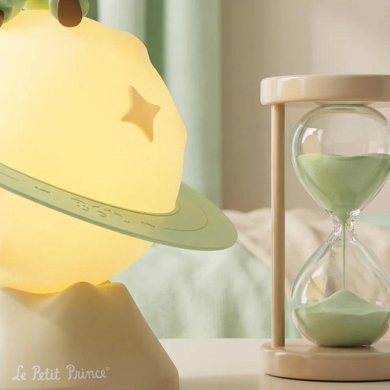 Plan macro montrant la texture de la Veilleuse Petit Prince jaune lumineuse avec une étoile et un anneau vert, posée à côté d'un sablier vert clair, idéale pour une chambre bébé.