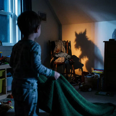 Enfant debout dans sa chambre sombre tenant une couverture, regardant l'ombre effrayante d'une chaise qui ressemble à un loup sur le mur. Illustration de la peur du noir et de l'imagination débordante chez les petits.