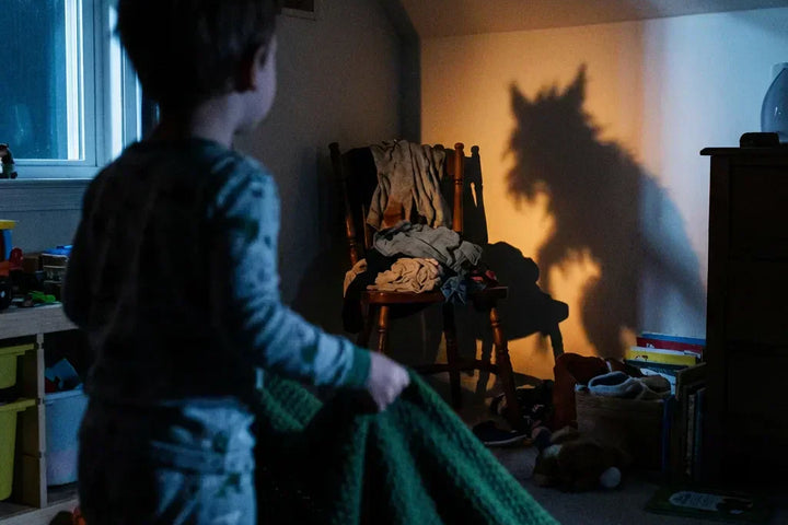 Enfant debout dans sa chambre sombre tenant une couverture, regardant l'ombre effrayante d'une chaise qui ressemble à un loup sur le mur. Illustration de la peur du noir et de l'imagination débordante chez les petits.