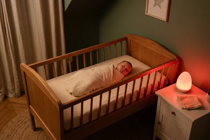 Rituel du coucher bébé sous lumière rouge 631 nm dans une chambre apaisée, nourrisson somnolent dans son lit en bois, veilleuse rouge allumée — Tendre Veilleuse