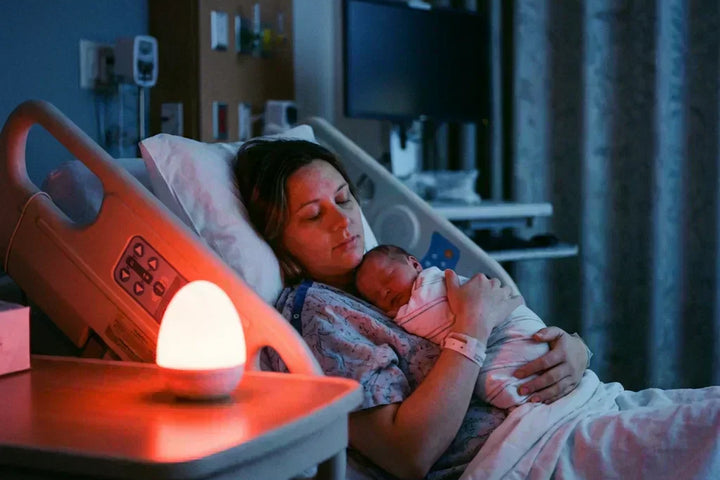 Maman et nouveau-né dormant paisiblement à l'hôpital, éclairés par la lumière rouge douce d'une veilleuse maternité nomade, idéale comme veilleuse allaitement la nuit.