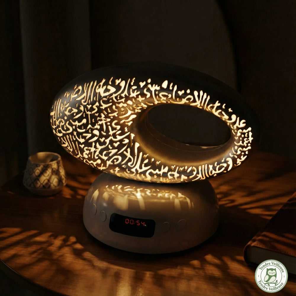 Vue nocturne du diffuseur coranique Souffle d'Orient illuminé, projetant des ombres de calligraphie arabe sur une table en bois avec horloge digitale visible.