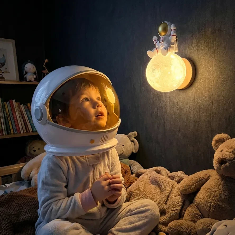 jeune enfant portant un grand casque d'astronaute, assis dans son lit entouré de peluches, levant les yeux avec émerveillement vers la veilleuse murale astronaute lune allumée sur un mur sombre.