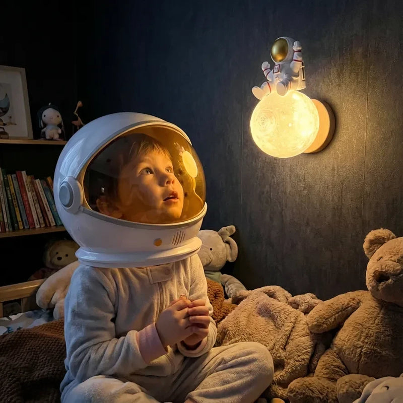 eune enfant portant un grand casque d'astronaute, assis dans son lit entouré de peluches, levant les yeux avec émerveillement vers la veilleuse murale astronaute lune allumée sur un mur sombre.