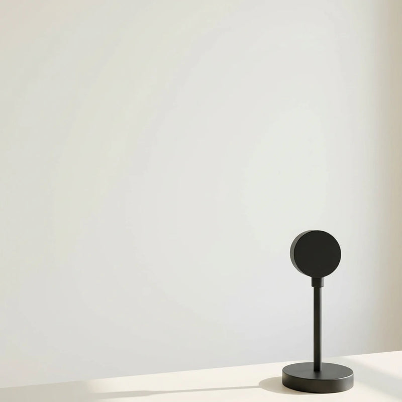 Lampe projecteur coucher de soleil noire éteinte sur un support, posée sur une surface blanche avec un fond blanc épuré.