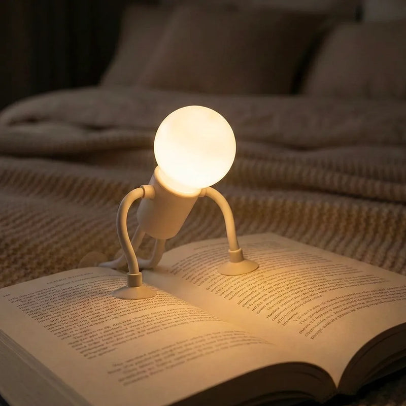 Lampe de lecture robot blanche posée en équilibre sur un livre ouvert dans un lit. Cette petite liseuse pour enfant diffuse une lumière chaude focalisée, idéale pour lire l'histoire du soir sans abîmer les yeux.