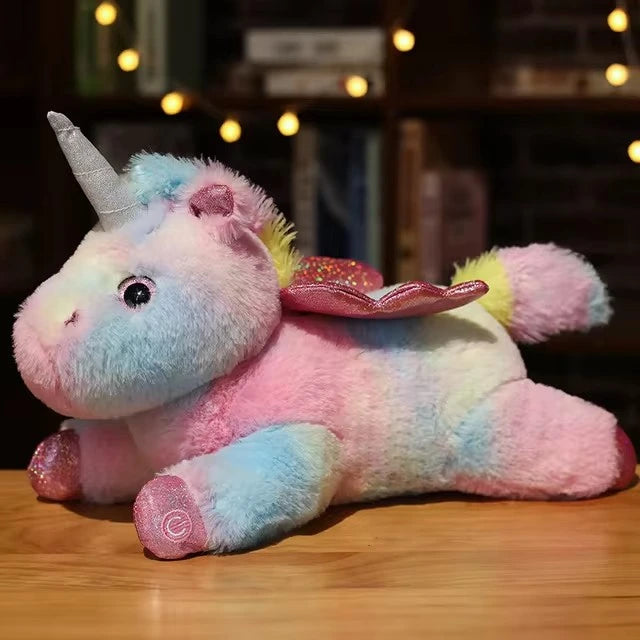 Peluche licorne aux couleurs arc-en-ciel pastel mélangées (rose, bleu, jaune) en position couchée, avec des ailes pailletées roses distinctives.