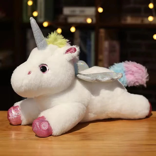 Peluche veilleuse licorne blanche en position couchée sur une table en bois, avec ailes argentées, crinière pastel et bouton d'allumage visible sur la patte.