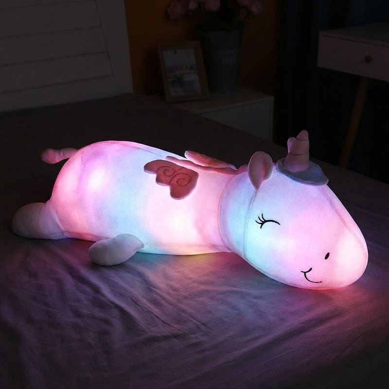 Peluche licorne géante illuminée de l'intérieur diffusant une douce lumière rose et violette dans une chambre sombre.
