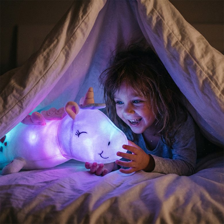 Enfant souriant caché sous une tente de draps, le visage éclairé par la lueur violette et magique d'une peluche licorne lumineuse.