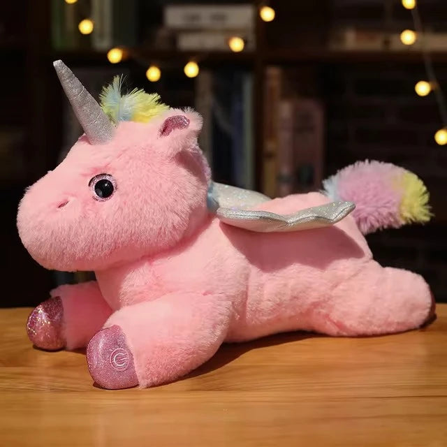 Peluche veilleuse licorne rose tendre en position allongée sur une surface boisée, affichant ses ailes argentées et sa crinière multicolore.