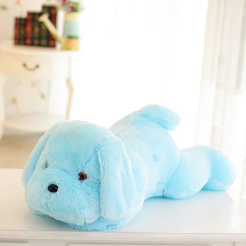Peluche chien lumineuse bleu ciel en position allongée sur une surface blanche, idéale comme coussin veilleuse doux pour chambre d'enfant.