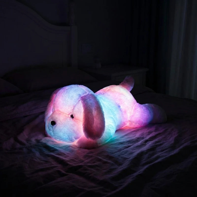 Peluche veilleuse chien rose illuminée de LED multicolores douces (rose, bleu, violet) dans une chambre sombre, créant une ambiance apaisante pour dormir.