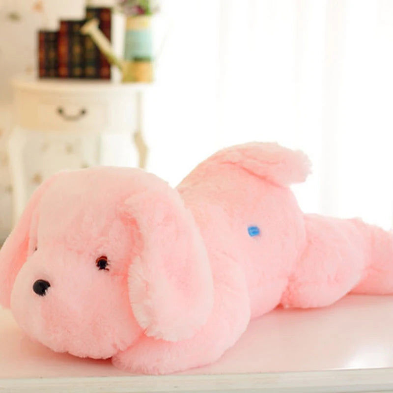 Vue de profil de la peluche chien rose allongée sur fond blanc, montrant son épaisseur moelleuse et le petit bouton d'activation bleu sur le flanc.