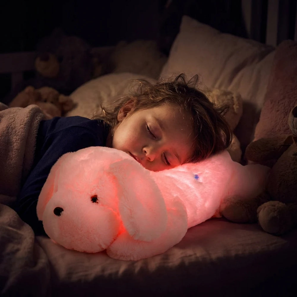 Enfant dormant profondément la tête posée sur sa peluche chien lumineuse rose, diffusant une lumière rassurante pour éviter la peur du noir.