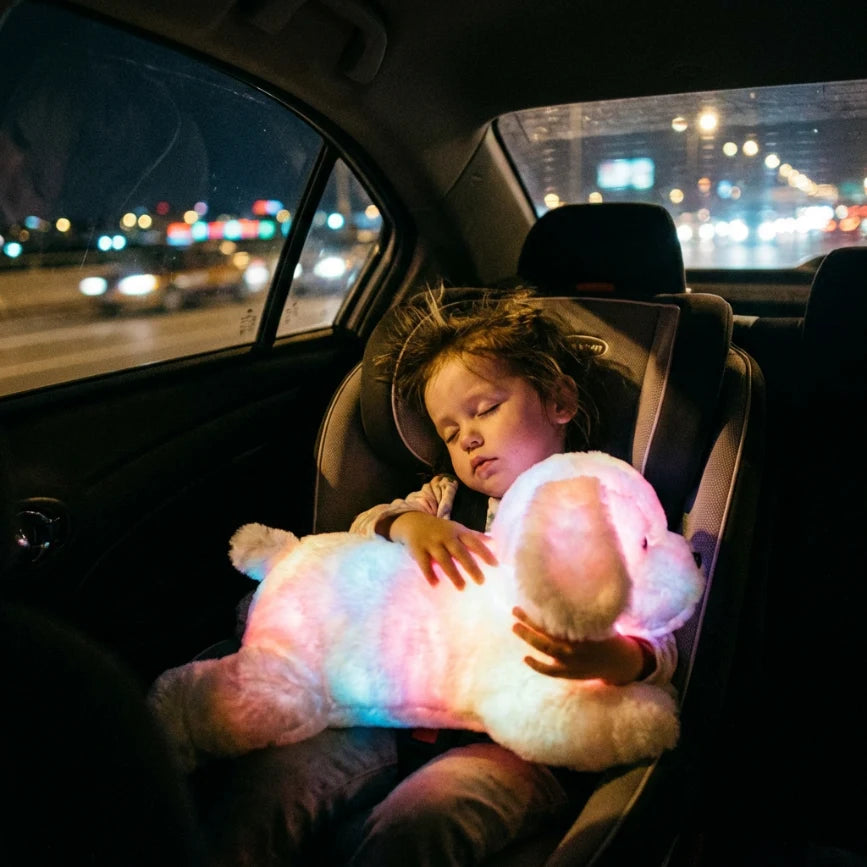 Enfant dormant paisiblement dans son siège auto, serrant contre lui sa peluche lumineuse chien blanche qui sert de veilleuse rassurante pour le voyage.