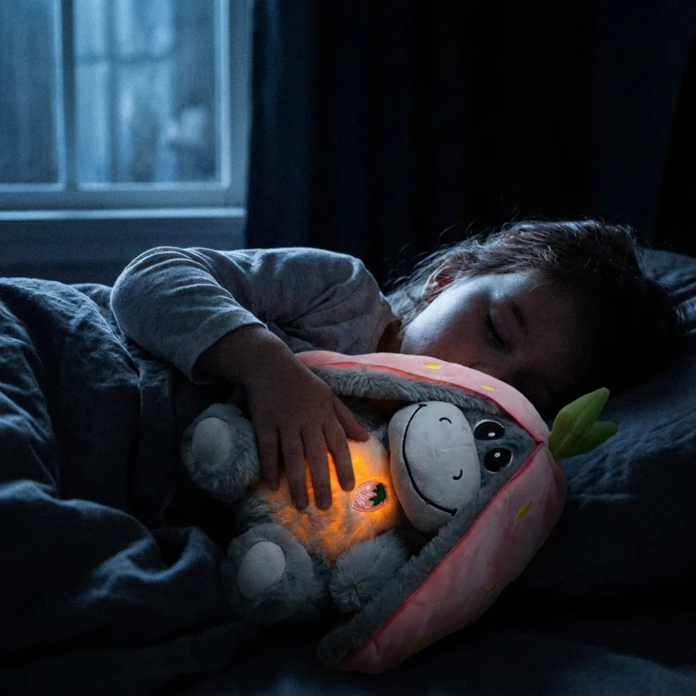 Enfant dormant profondément dans la pénombre, serrant contre lui la peluche âne fraise pour s'apaiser grâce à son rythme respiratoire et sa douceur.