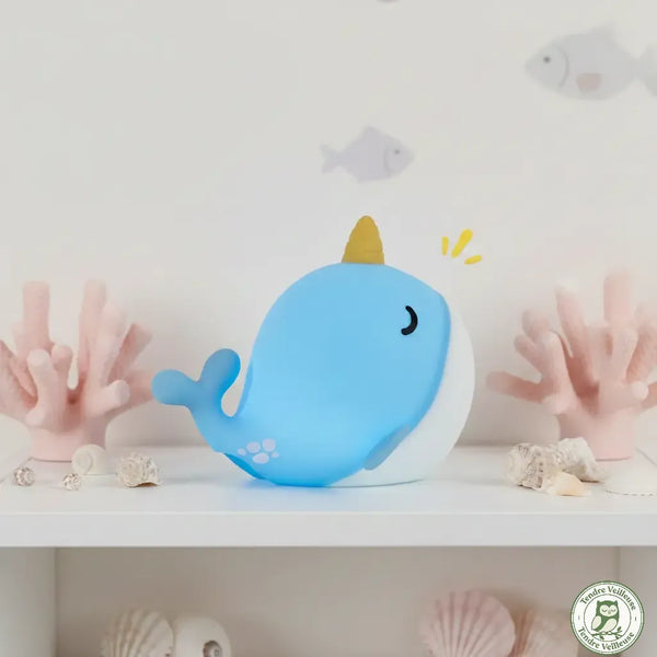 Veilleuse baleine bleue et blanche en silicone souple posée sur une étagère déco marine, avec corne dorée façon licorne