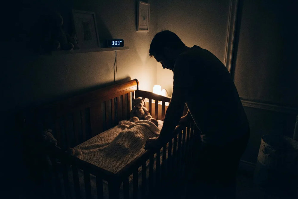 Un papa debout près du berceau de son bébé endormi à 3h03 du matin dans une chambre sombre. Une veilleuse nomade à lumière chaude diffuse une lueur rassurante pour apaiser l'angoisse de séparation et la peur du noir du nouveau-né.