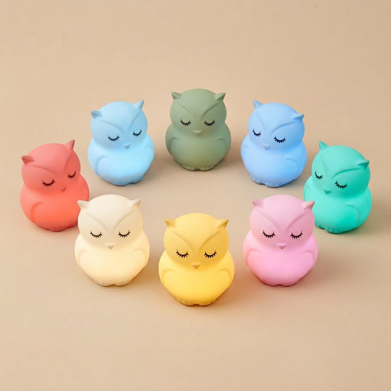 Plusieurs veilleuses hibou silicone Noctou alignées en arc, chacune allumée dans une couleur différente — blanc, rouge, vert, bleu, jaune, rose, cyan — fond beige Tendre Veilleuse
