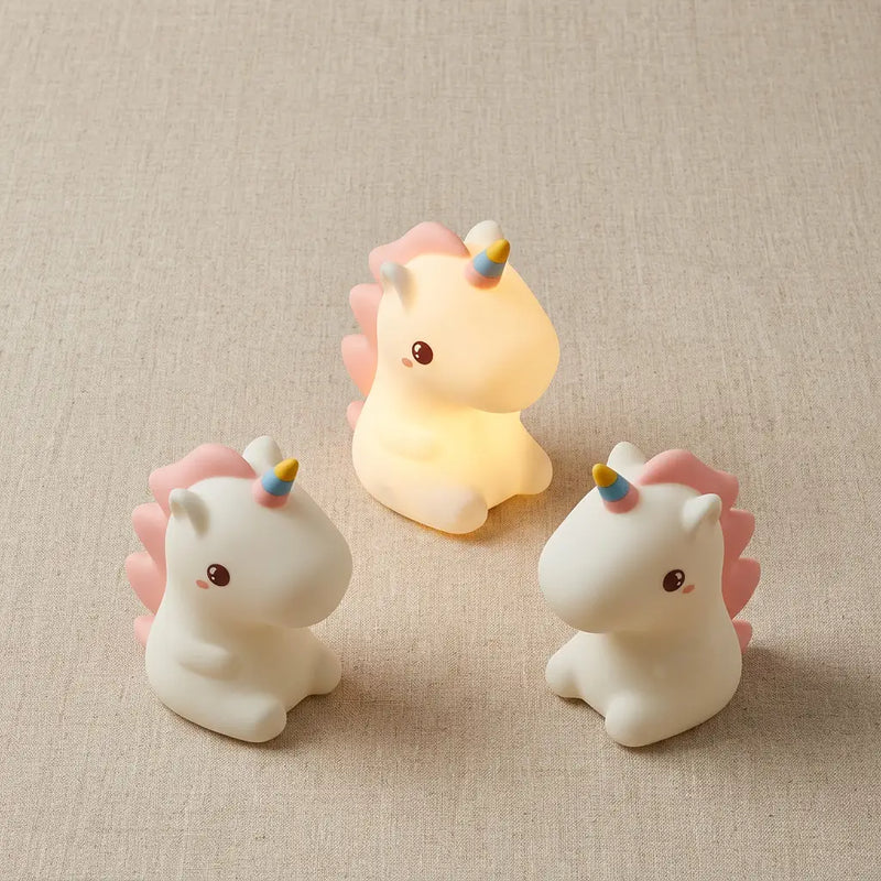 Trois veilleuses licorne silicone disposées en flat lay sur tissu lin naturel, une allumée avec halo de lumière douce, deux éteintes. Tendre Veilleuse