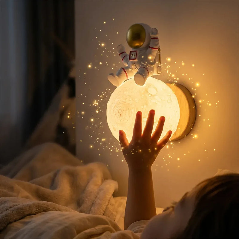 La main d'un jeune enfant tendue vers la veilleuse murale astronaute lune allumée, diffusant une lumière chaude et féérique dans une chambre sombre.