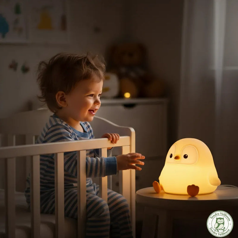 Bébé en pyjama rayé souriant vers veilleuse pingouin Pongo dans sa chambre la nuit, lumière chaude douce sommeil rechargeable, Tendre Veilleuse