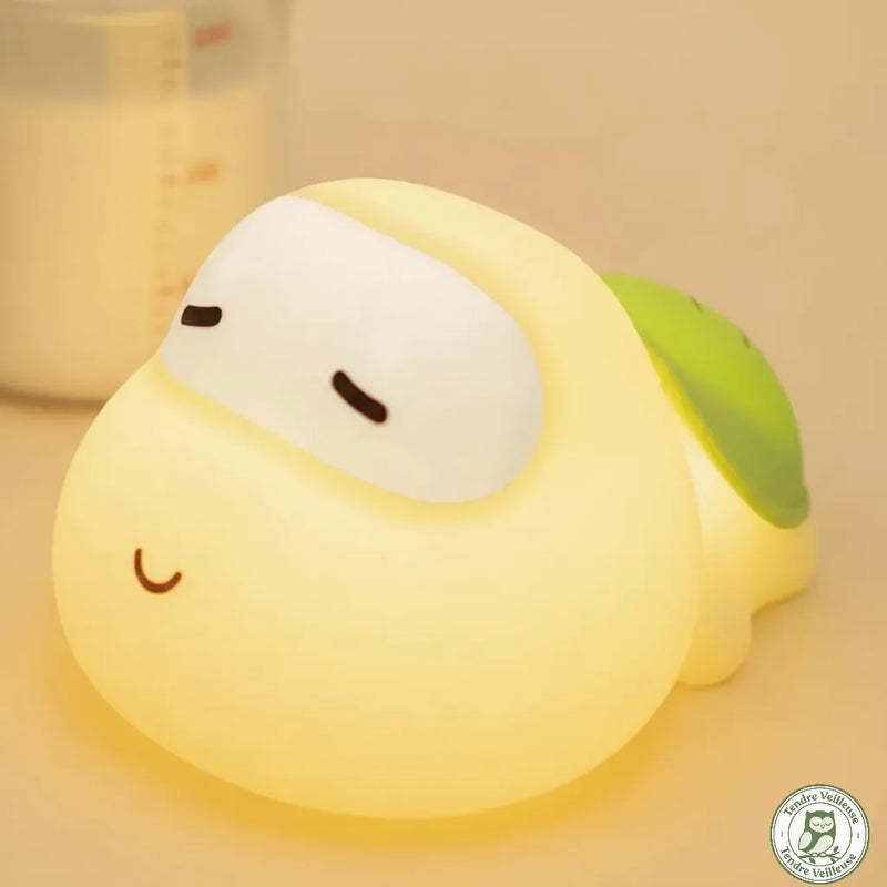 Gros plan veilleuse tortue bébé silicone Titou allumée lumière chaude jaune, biberon visible en arrière-plan, usage allaitement nocturne — Tendre Veilleuse
